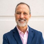 A smiling man with a grey beard and wearing a blue suit